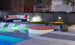 Relax on this modern rooftop terrace with a pool featuring submerged lounge chairs, a Ledge Lounger Signature Round Sunbed, striped sofa with red pillows, ambient lighting, and lush greenery. Enjoy the perfect cityscape backdrop for unwinding.