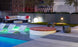 Relax on this modern rooftop terrace with a pool featuring submerged lounge chairs, a Ledge Lounger Signature Round Sunbed, striped sofa with red pillows, ambient lighting, and lush greenery. Enjoy the perfect cityscape backdrop for unwinding.