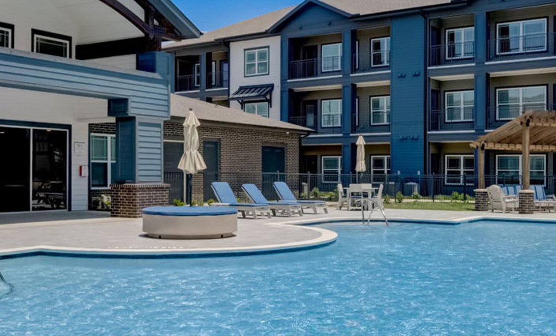 Ledge Loungers Signature Round Sunbed invites relaxation by the outdoor pool of a modern three-story apartment with blue and white accents, complemented by lounge chairs, umbrellas, and a circular hot tub.