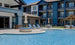 Ledge Loungers Signature Round Sunbed invites relaxation by the outdoor pool of a modern three-story apartment with blue and white accents, complemented by lounge chairs, umbrellas, and a circular hot tub.
