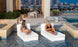 Two women relax on Ledge Loungers Signature Sectional 2 Piece Sun Chairs, partially submerged in a pool. They sit across from each other with modern architecture and a nighttime cityscape in the background, while tables and chairs on the patio complete the serene scene.