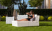 A woman in a black dress relaxes on a Signature Sectional 2 Piece Sun Chair by Ledge Lounger, surrounded by lush green plants and distant buildings. Wearing sunglasses and holding her smartphone, she enjoys the serene garden atmosphere.
