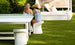 Two women relax at a modern white outdoor table on a green lawn. One wears a blue dress, the other a blue and white swimsuit with a hat. Nearby, stylish Signature Barstool Counter Height chairs by Ledge Lounger complement the elegant setting for their sunny garden day.