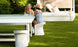 Two women relax at a modern white outdoor table on a green lawn. One wears a blue dress, the other a blue and white swimsuit with a hat. Nearby, stylish Signature Barstool Counter Height chairs by Ledge Lounger complement the elegant setting for their sunny garden day.