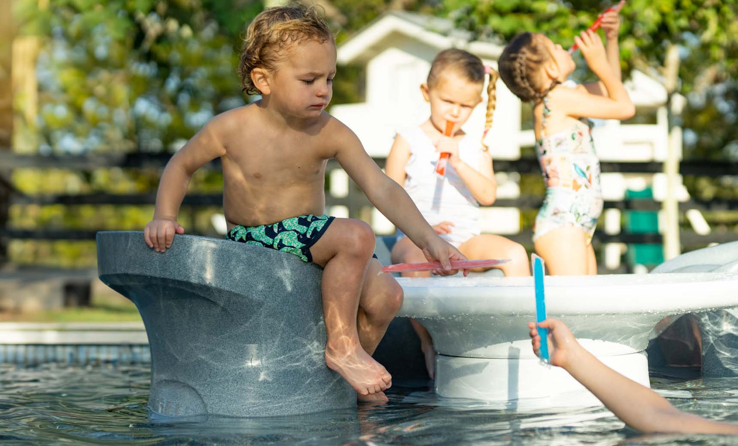 Under the sun, three children savor ice pops by a pool. One sits at the edge, another stands close, and the third drinks from a straw. In the foreground, near some Ledge Lounger Signature Barstool Counter Height seats beneath the water, a hand holding an ice pop appears.