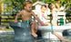 Under the sun, three children savor ice pops by a pool. One sits at the edge, another stands close, and the third drinks from a straw. In the foreground, near some Ledge Lounger Signature Barstool Counter Height seats beneath the water, a hand holding an ice pop appears.