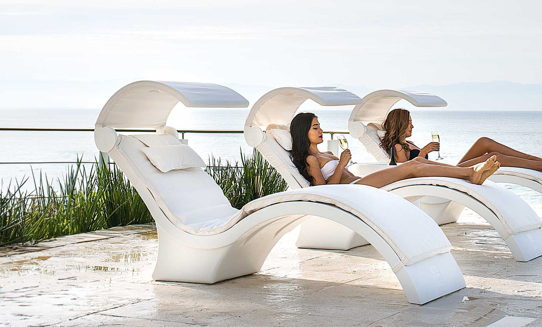 Two people relax on Ledge Lounger’s Signature Chaise Deep Cushion lounges with built-in canopies by a pool, overlooking the ocean. Wearing swimsuits and holding glasses, they enjoy a serene, sunny day in this tranquil and luxurious setting.