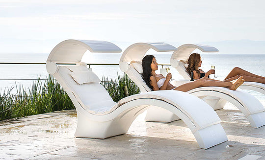 Two people relax on Ledge Lounger’s Signature Chaise Deep Cushion lounges with built-in canopies by a pool, overlooking the ocean. Wearing swimsuits and holding glasses, they enjoy a serene, sunny day in this tranquil and luxurious setting.