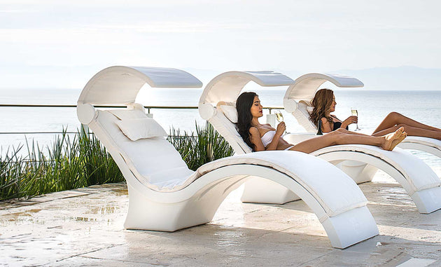 Two people relax on Ledge Lounger’s Signature Chaise Deep Cushion lounges with built-in canopies by a pool, overlooking the ocean. Wearing swimsuits and holding glasses, they enjoy a serene, sunny day in this tranquil and luxurious setting.