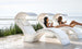 Two people relax on Ledge Lounger’s Signature Chaise Deep Cushion lounges with built-in canopies by a pool, overlooking the ocean. Wearing swimsuits and holding glasses, they enjoy a serene, sunny day in this tranquil and luxurious setting.