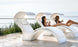 Two people relax on Ledge Lounger’s Signature Chaise Deep Cushion lounges with built-in canopies by a pool, overlooking the ocean. Wearing swimsuits and holding glasses, they enjoy a serene, sunny day in this tranquil and luxurious setting.