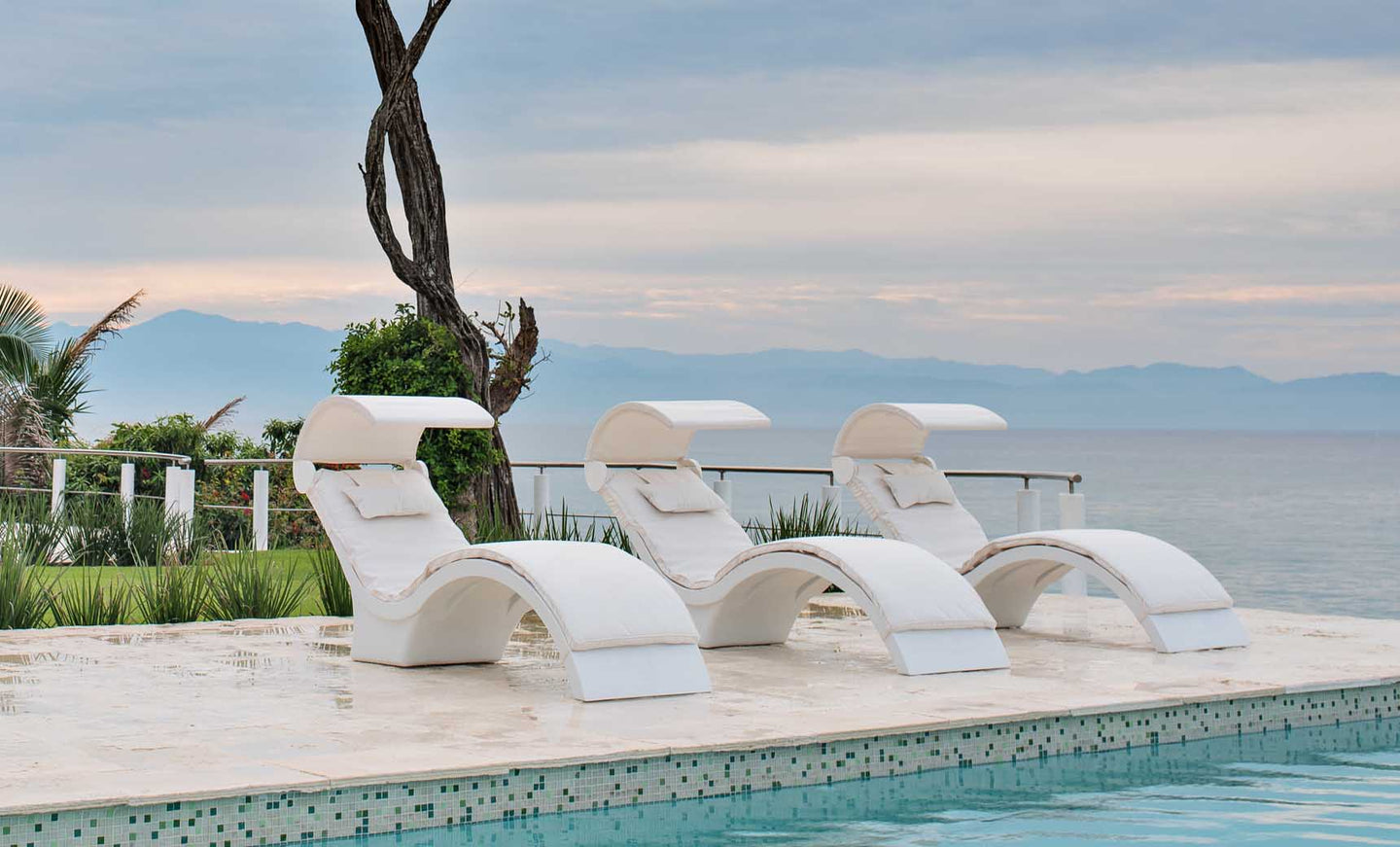 Three Signature Chaise Deep Cushions by Ledge Lounger are positioned beside a clear blue pool. In the background, enjoy a calm ocean view with distant mountains under a cloudy sky, complemented by greenery and a solitary tree to the left.