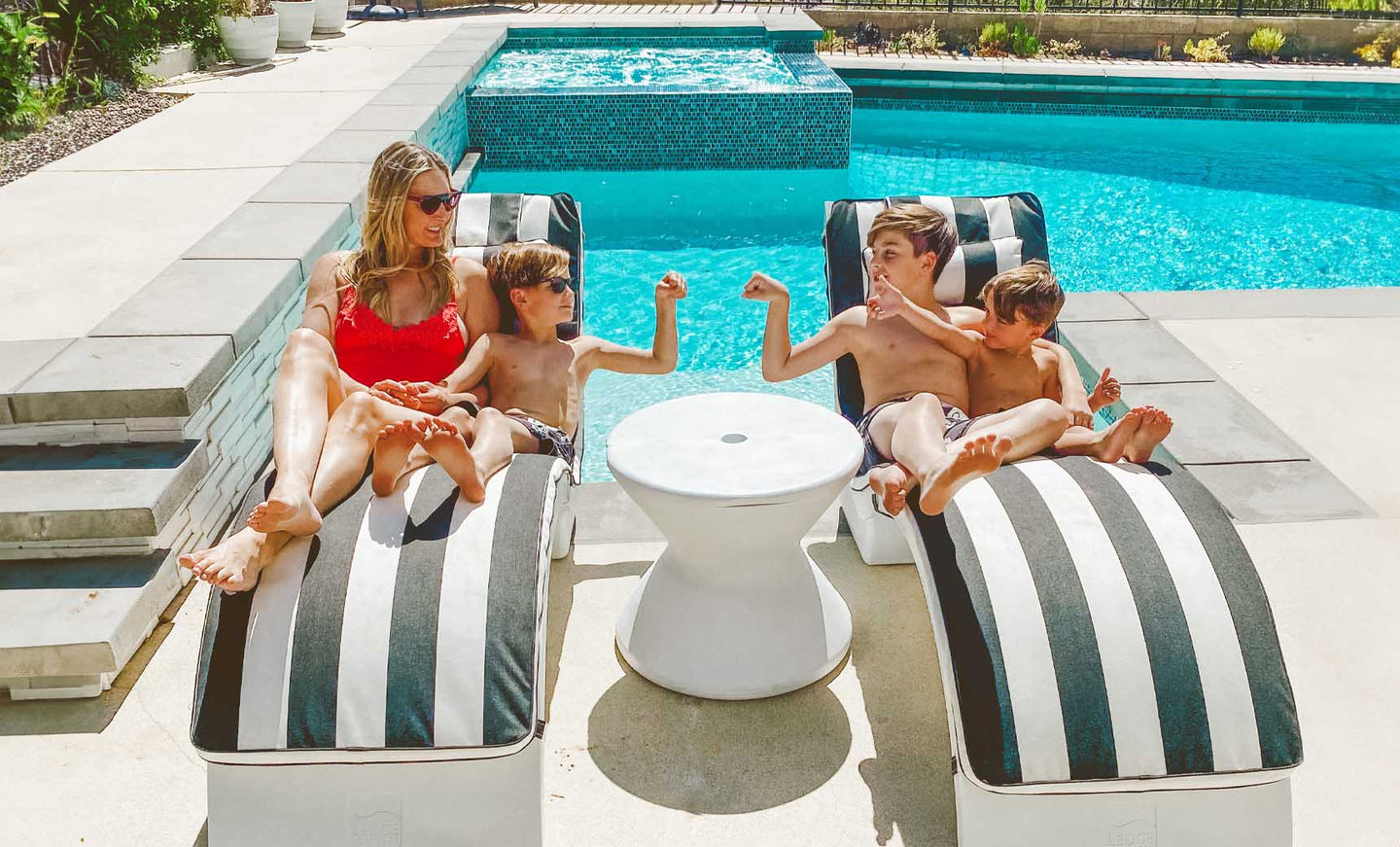 A woman in a red swimsuit and sunglasses relaxes by the pool on a Ledge Lounger Signature Chaise Deep Cushion. Three children are with her, one playfully flexing while the others enjoy the sun. A small round table sits between their striped loungers.