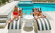 A woman in a red swimsuit and sunglasses relaxes by the pool on a Ledge Lounger Signature Chaise Deep Cushion. Three children are with her, one playfully flexing while the others enjoy the sun. A small round table sits between their striped loungers.
