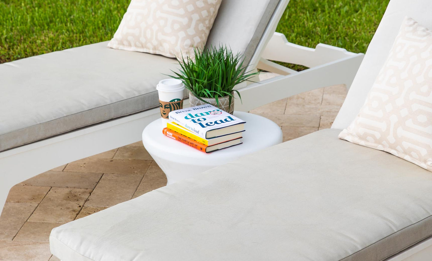 Two white lounge chairs with beige cushions and patterned pillows flank the Signature Standard Side Table by Ledge Lounger. The round table holds three books, a small plant, and a coffee cup, set against a backdrop of green grass.