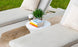 Two white lounge chairs with beige cushions and patterned pillows flank the Signature Standard Side Table by Ledge Lounger. The round table holds three books, a small plant, and a coffee cup, set against a backdrop of green grass.