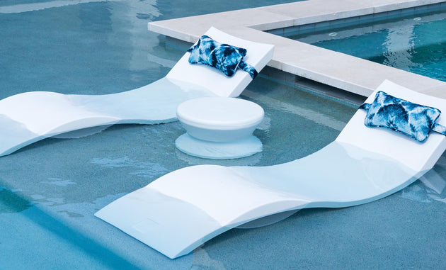 On a sun shelf, two modern white pool loungers with blue patterned pillows are partially submerged next to a Ledge Lounger Signature Standard Side Table, perfectly complementing the clear turquoise water.