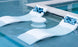 On a sun shelf, two modern white pool loungers with blue patterned pillows are partially submerged next to a Ledge Lounger Signature Standard Side Table, perfectly complementing the clear turquoise water.
