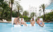 Four people enjoy drinks and lively conversation on Ledge Loungers Signature Chairs surrounded by palm trees. Two women and two men in swimsuits relax in the durable, comfortable loungers, with a large building in the background of this serene hotel poolside scene.