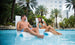 A man and woman relax on a Signature Chair from Ledge Lounger in a pool surrounded by palm trees. The man wears blue swim shorts, and the woman enjoys her drink in a blue patterned swimsuit, their cheerful conversation enhancing the serene atmosphere.