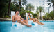 A man and woman relax on a Signature Chair from Ledge Lounger in a pool surrounded by palm trees. The man wears blue swim shorts, and the woman enjoys her drink in a blue patterned swimsuit, their cheerful conversation enhancing the serene atmosphere.