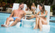 A man and woman in swimsuits relax on Ledge Lounger Signature Chairs partially submerged on a tanning ledge, smiling at each other over a round table with drinks and sunglasses. Two others recline on poolside chairs in the background, surrounded by lush greenery in this outdoor oasis.