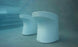 Two white Ledge Lounger Signature Barstools (Counter Height) sit elegantly at the bottom of a swimming pool. Their modern design stands out against the blue tiled steps in the background, with water creating a gentle blur effect.