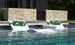 Two Ledge Lounger white chairs with green leaf towels offer in-pool relaxation, submerged in water. Between them, a round white table awaits customizable options like a refreshing drink. The setting is complemented by a stone wall and lush greenery. Signature Headrest Pillow adds extra comfort.