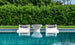 Two white Ledge Lounger Signature Headrest Pillow loungers with gray cushions rest partly submerged in a clear blue pool, flanking a small white table. Lush green foliage forms a dense background, enhancing the natural tranquility of this in-pool relaxation setting.