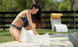 A woman in a blue bikini assembles a white Signature Chaise Deep Riser by the poolside on an outdoor day with other Ledge Lounger in-pool seats, lush grass, swaying trees, and a charming wooden fence in the backdrop.