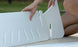 Someone is assembling Ledge Loungers Signature Chaise Riser on a wooden surface, aligning two white plastic panels with slotted vents and text. One hand, visible with a ring, is shown. The background features a blurred outdoor scene with grass.