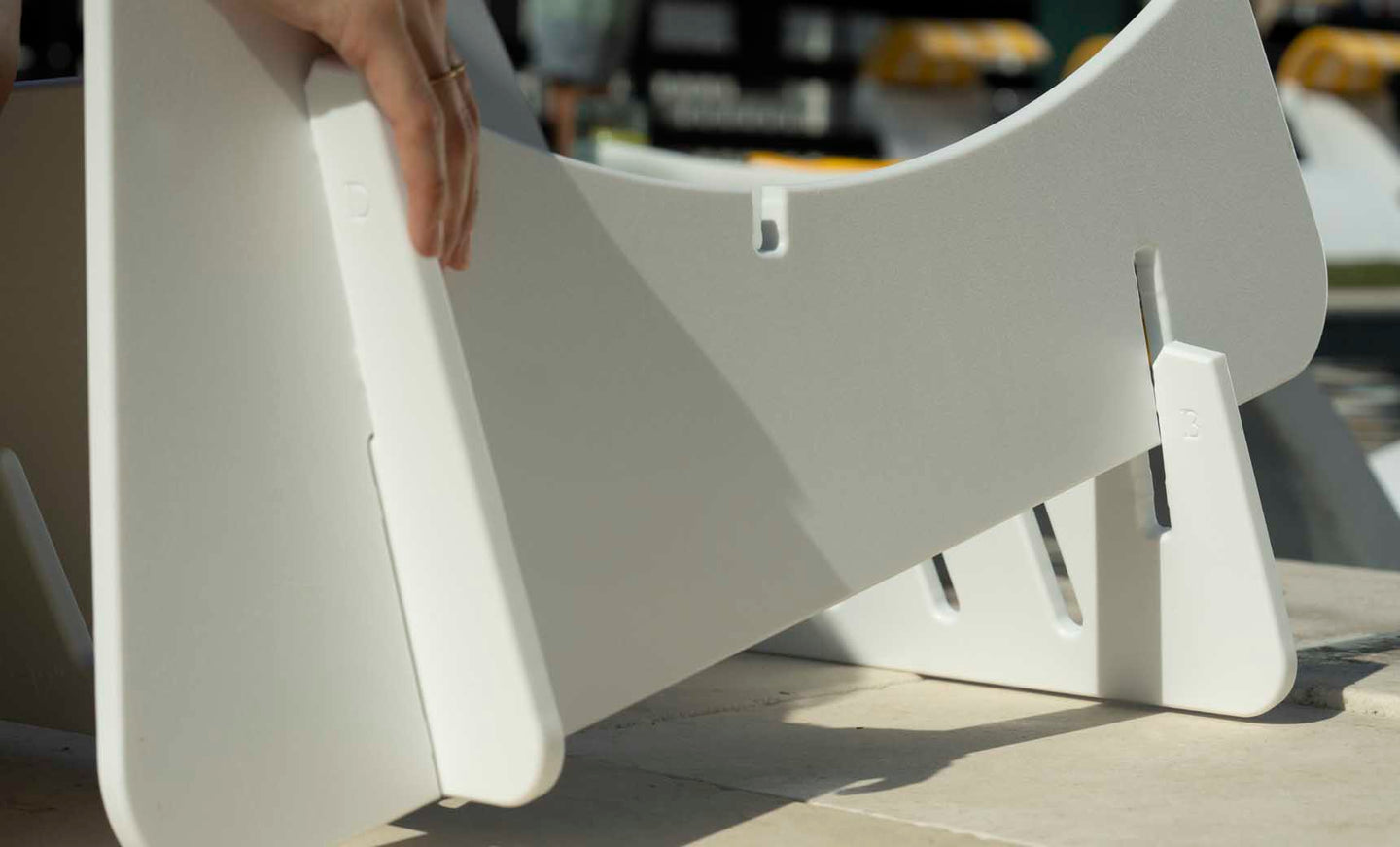 A person skillfully assembles a white, modular Ledge Lounger Signature Chair Riser indoors, precisely fitting two slotted parts together. The soft shadow on the light surface highlights the risers elegance.