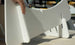 A person skillfully assembles a white, modular Ledge Lounger Signature Chair Riser indoors, precisely fitting two slotted parts together. The soft shadow on the light surface highlights the risers elegance.