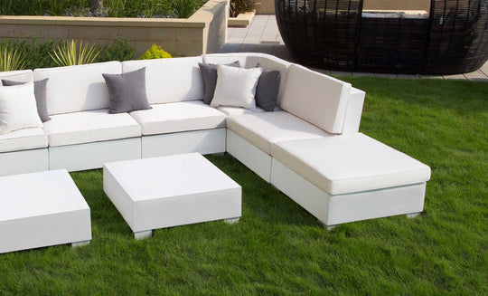 A Signature Sectional Corner by Ledge Lounger, featuring gray and white pillows, sits on green grass. Two white square tables add elegance in front. A round wicker chair rests on the paved area behind, completing this serene setting.