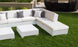A Signature Sectional Corner by Ledge Lounger, featuring gray and white pillows, sits on green grass. Two white square tables add elegance in front. A round wicker chair rests on the paved area behind, completing this serene setting.