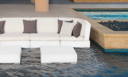 A modern outdoor seating area highlights Ledge Loungers Signature Sectional Corner couches with white cushions and dark pillows partially submerged. Two square, white tables sit in front, near a black-pebbled pool with beige stone borders.