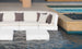 A modern outdoor seating area highlights Ledge Loungers Signature Sectional Corner couches with white cushions and dark pillows partially submerged. Two square, white tables sit in front, near a black-pebbled pool with beige stone borders.