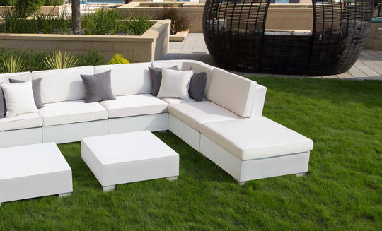 An outdoor lounge area featuring a Ledge Lounger Signature Sectional Ottoman and white sectional sofas sits on a grassy lawn. The seating is decorated with gray and white pillows, while a large round wicker structure and landscaped planters enhance the background.