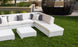 An outdoor lounge area featuring a Ledge Lounger Signature Sectional Ottoman and white sectional sofas sits on a grassy lawn. The seating is decorated with gray and white pillows, while a large round wicker structure and landscaped planters enhance the background.