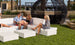 People relax on white outdoor couches atop a grassy rooftop with modern furniture. A large black spherical structure looms in the background against a cityscape, while the Ledge Lounger Signature Sectional Ottoman with its sleek one-piece base and plush cushion adds style and comfort.