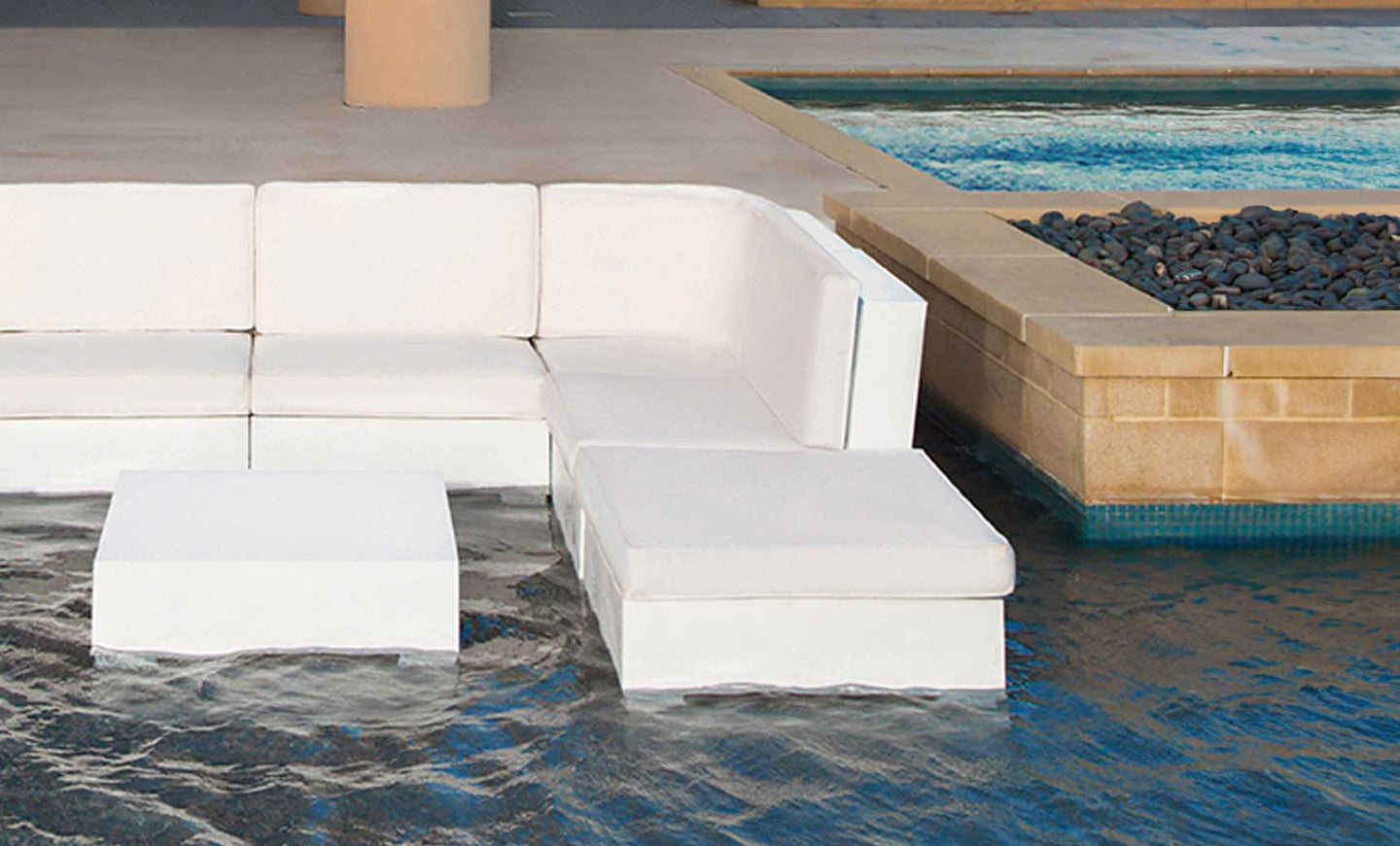 A Ledge Lounger Signature Sectional Ottoman with cushions and white outdoor modular seating are partially submerged in a pool, next to a tiled ledge decorated with black stones.