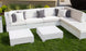 The outdoor patio features the Ledge Lounger Signature Sectional Middle with a white modular design and two-piece cushions. Gray and white throw pillows add elegance, while matching white square tables rest on green grass. Lush plants and a stone wall create a serene background.