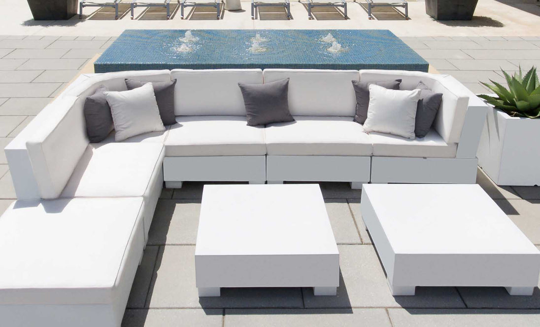 The patio set showcases a Ledge Lounger Signature Sectional Middle with white sofas and two-piece cushions, highlighted by gray pillows. Nearby are two square white tables, backed by a small fountain on tiles, while lounge chairs rest in the distance.