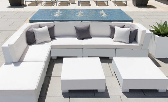 The patio set showcases a Ledge Lounger Signature Sectional Middle with white sofas and two-piece cushions, highlighted by gray pillows. Nearby are two square white tables, backed by a small fountain on tiles, while lounge chairs rest in the distance.