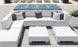 The patio set showcases a Ledge Lounger Signature Sectional Middle with white sofas and two-piece cushions, highlighted by gray pillows. Nearby are two square white tables, backed by a small fountain on tiles, while lounge chairs rest in the distance.