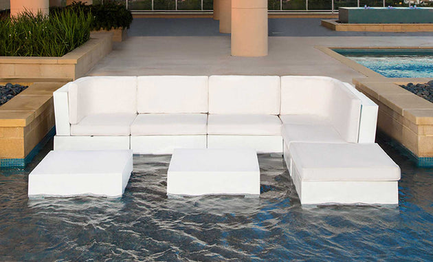 The Signature Sectional Middle by Ledge Lounger, featuring two-piece cushions, offers luxury outdoors. This modern white sectional and three tables are partly submerged in a shallow pool, with water rippling gently, enhancing the tranquility near planted areas and the larger swimming pool.