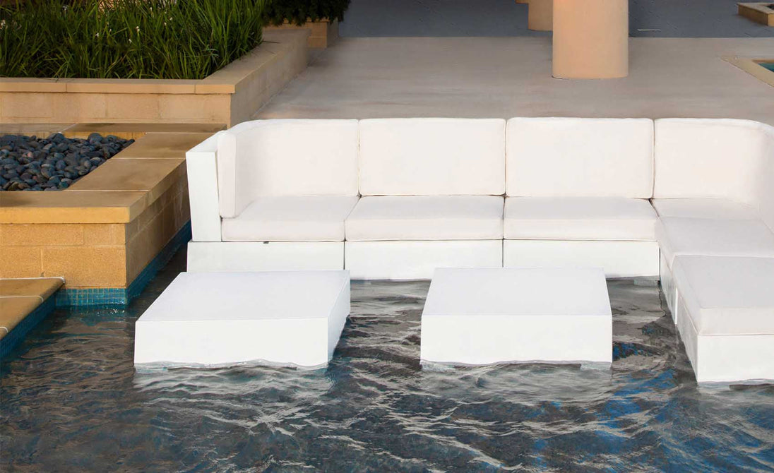 A modern outdoor area features a white Ledge Lounger Signature Sectional Endcap partially submerged in a shallow pool with ottomans. Two square white tables sit at the corners, surrounded by stone walls and elegant potted plants.