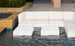 A modern outdoor area features a white Ledge Lounger Signature Sectional Endcap partially submerged in a shallow pool with ottomans. Two square white tables sit at the corners, surrounded by stone walls and elegant potted plants.