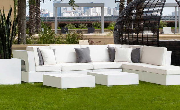 The Signature Sectional Endcap by Ledge Lounger, with gray and white cushions, sits on green grass alongside elegant coffee tables. A large wicker chair is nearby, and the city skyline is framed by plants and palm trees adorning the terrace.
