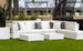 The Signature Sectional Endcap by Ledge Lounger, with gray and white cushions, sits on green grass alongside elegant coffee tables. A large wicker chair is nearby, and the city skyline is framed by plants and palm trees adorning the terrace.